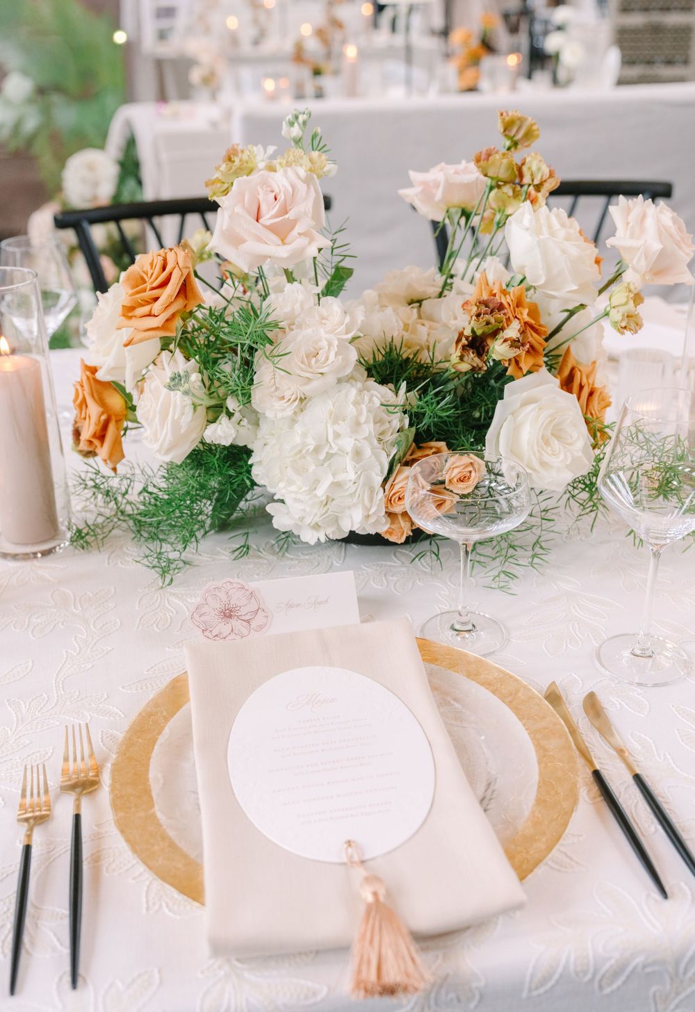 Elegant wedding table setting with gold charger plates, black cutlery, circular menu card, and peach and white floral centerpiece.