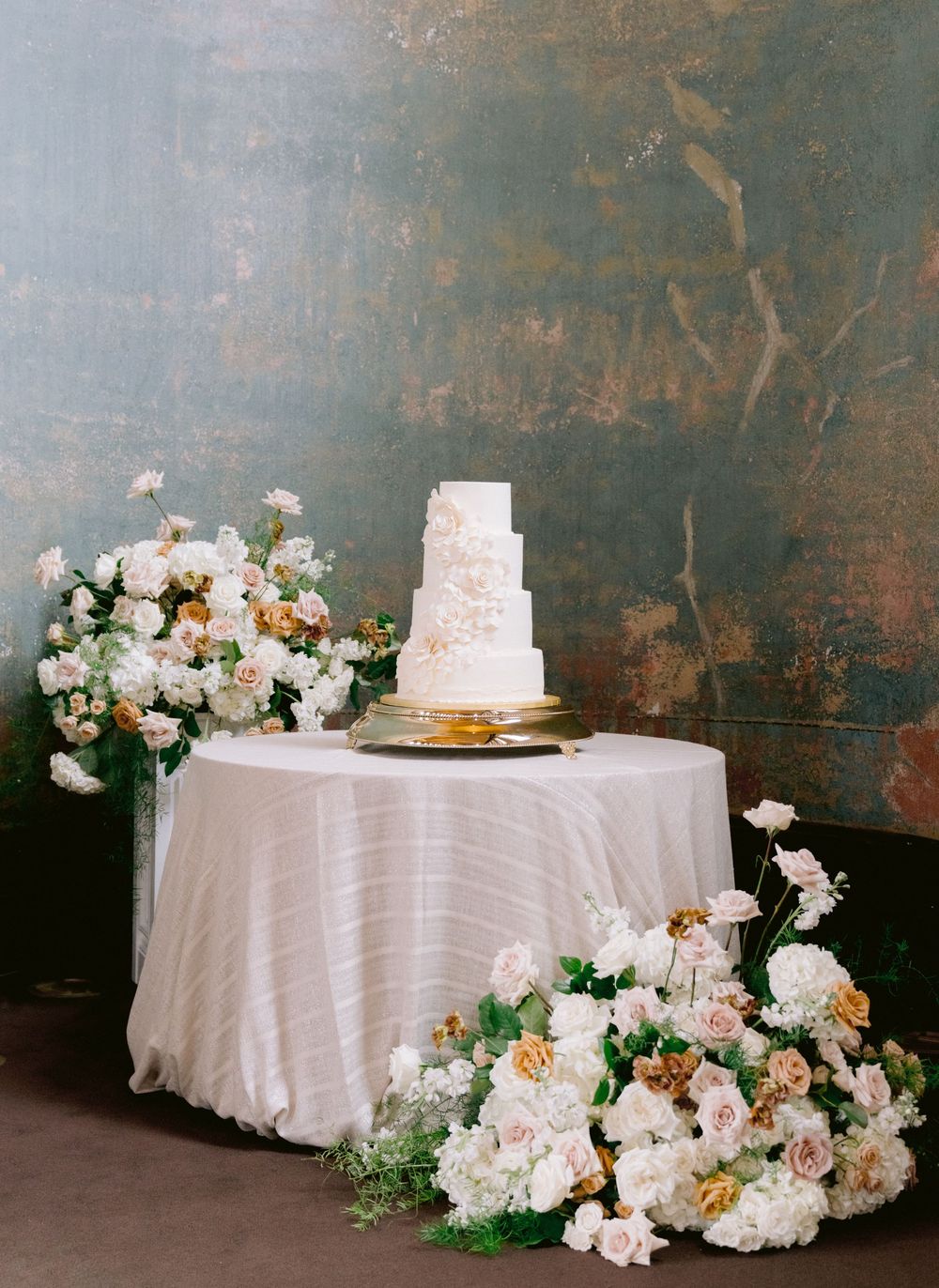 Elegant four-tier white wedding cake on a draped table surrounded by lush floral arrangements of white and blush roses.