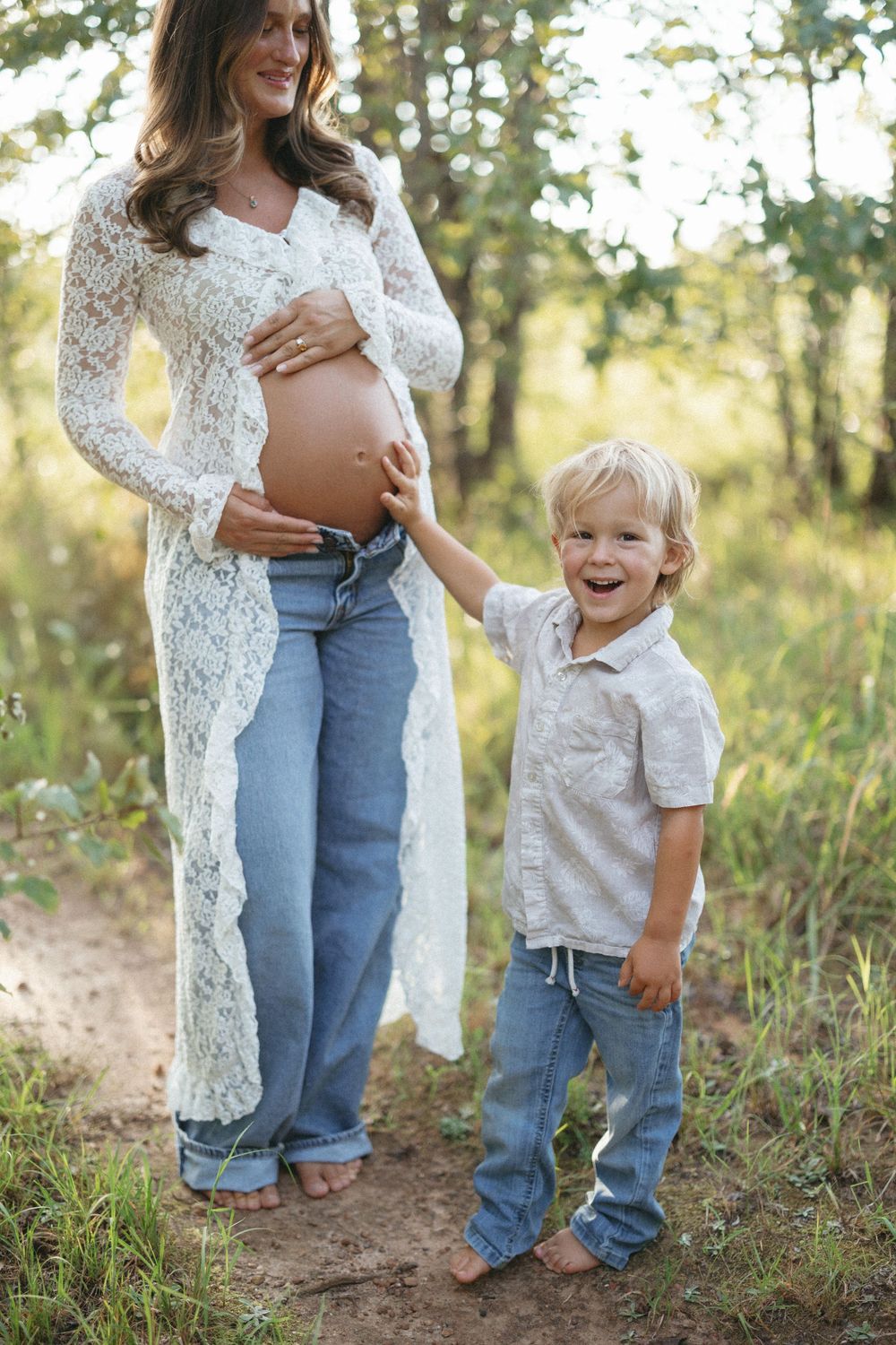 Smiling toddler boy touches pregnant mothers bare belly outdoors, both wearing jeans in sunny nature setting.