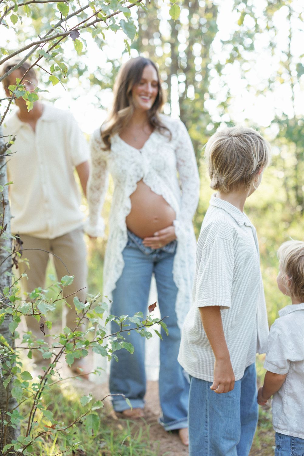 Pregnant woman in lace top and jeans holds belly outdoors, smiling at two young boys in a sunlit forest setting.