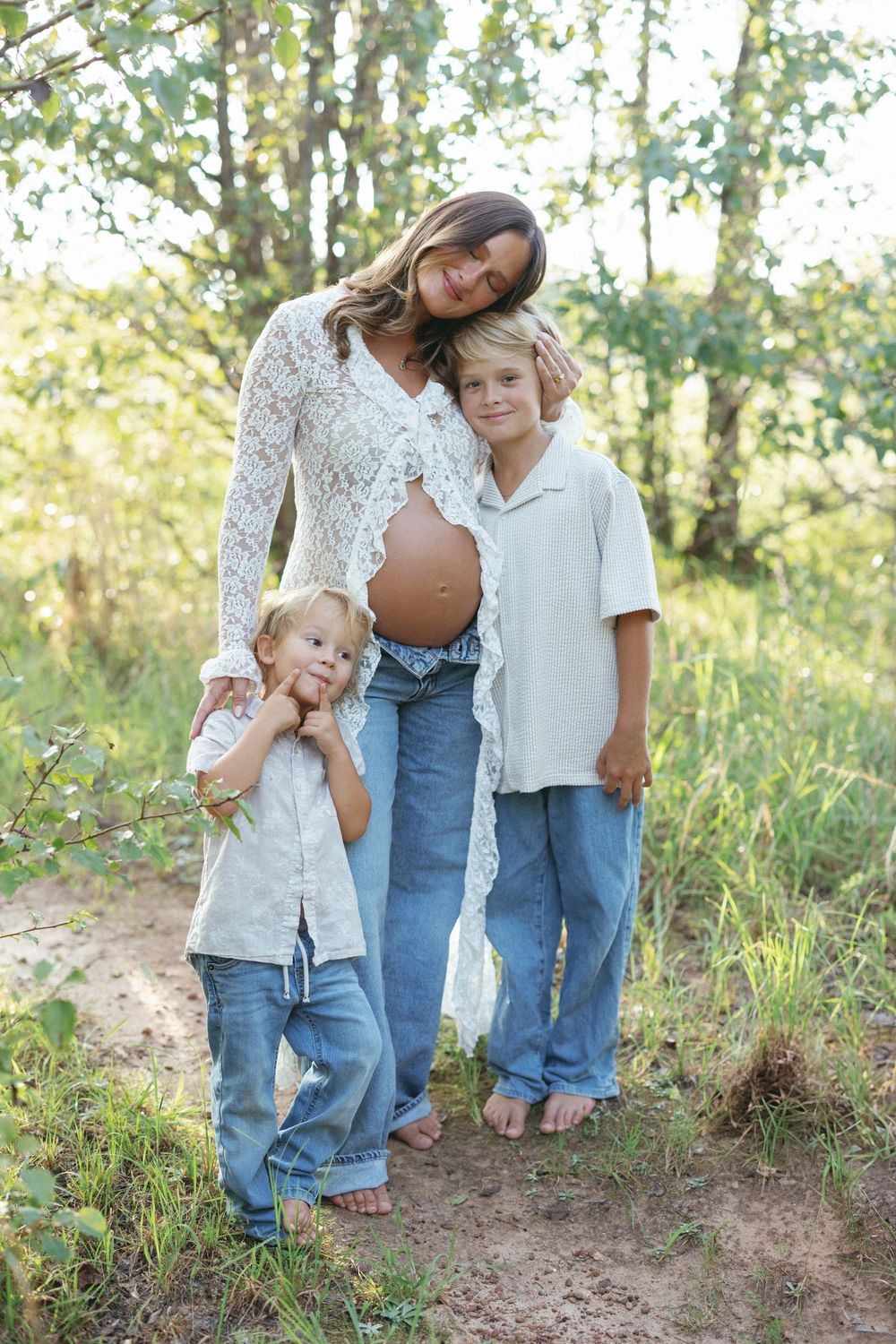 Pregnant woman in lace cardigan and jeans poses outdoors with two young sons in a sunny green forest setting.