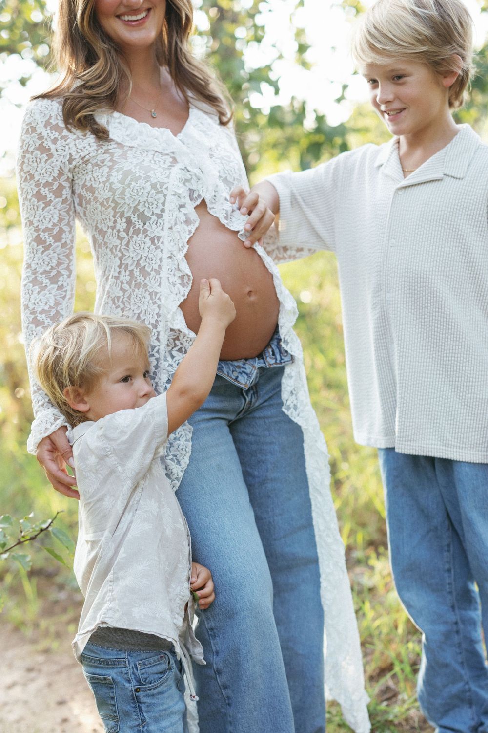Pregnant woman in lace overlay and jeans outdoors with two young blonde boys touching her baby bump.