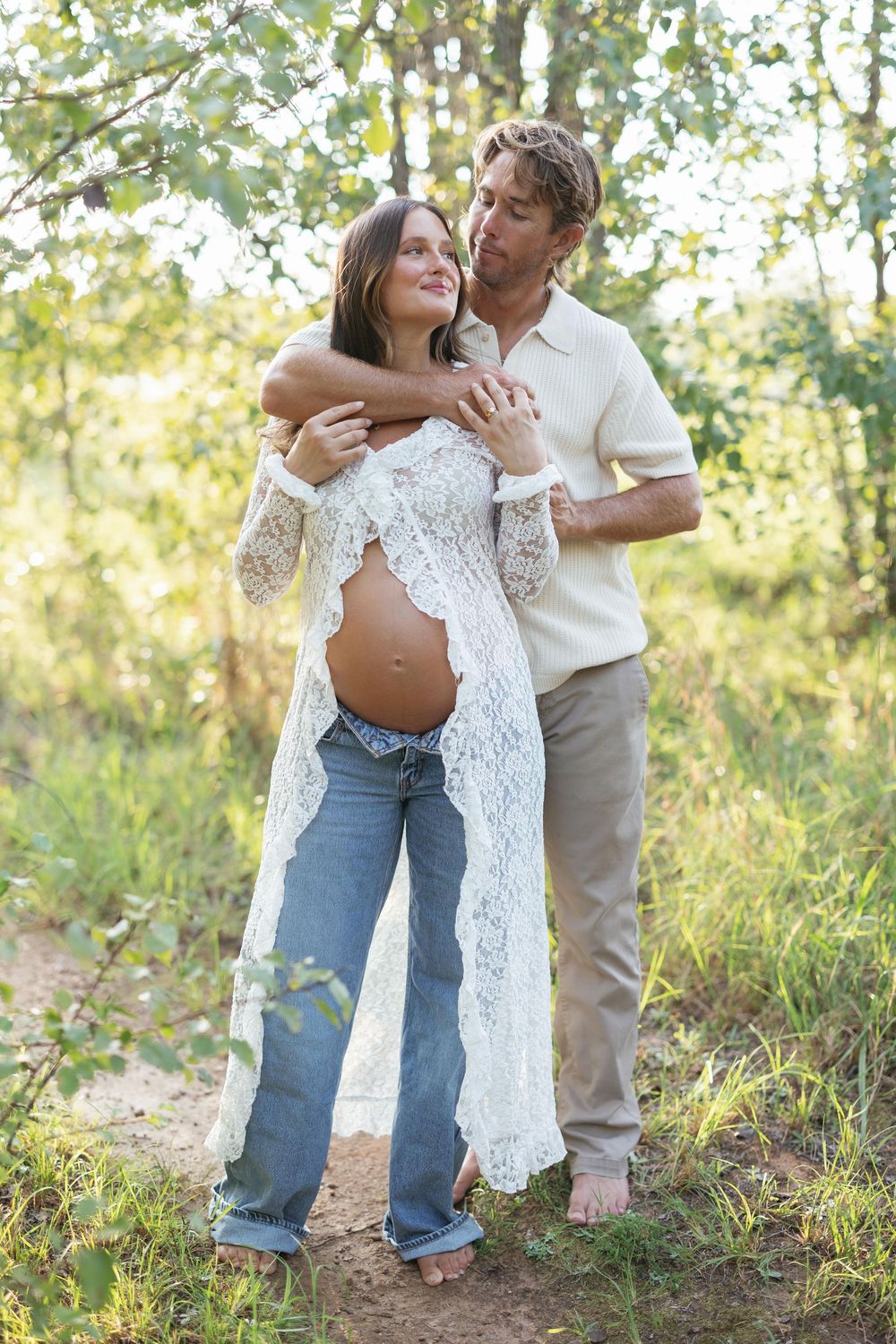 Couple in nature, pregnant woman in white lace cardigan and jeans embraced by partner among green trees.