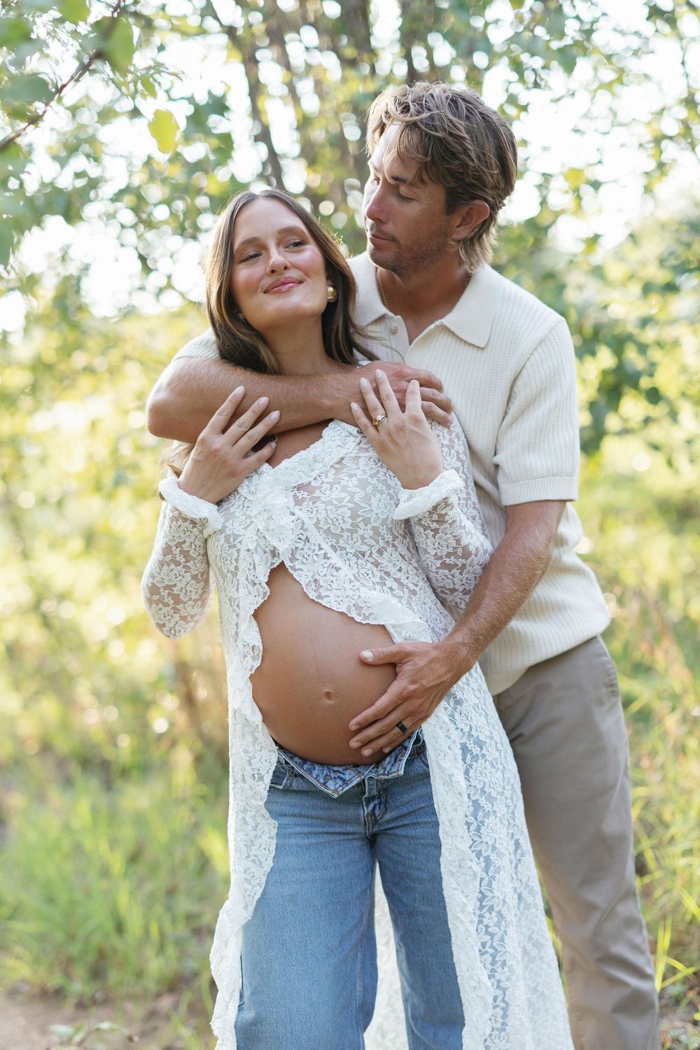 Couple sharing tender maternity moment outdoors, man embracing pregnant woman in white lace.