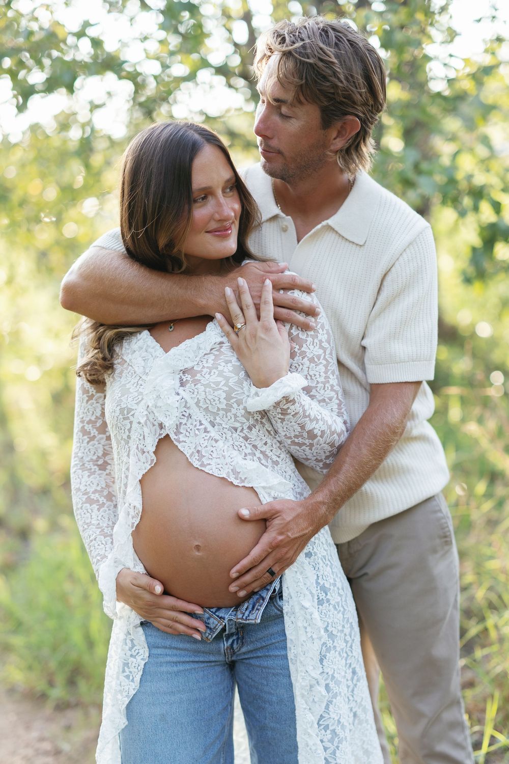 Couple sharing tender moment outdoors, pregnant woman in lace top and jeans cradling baby bump while partner embraces her.