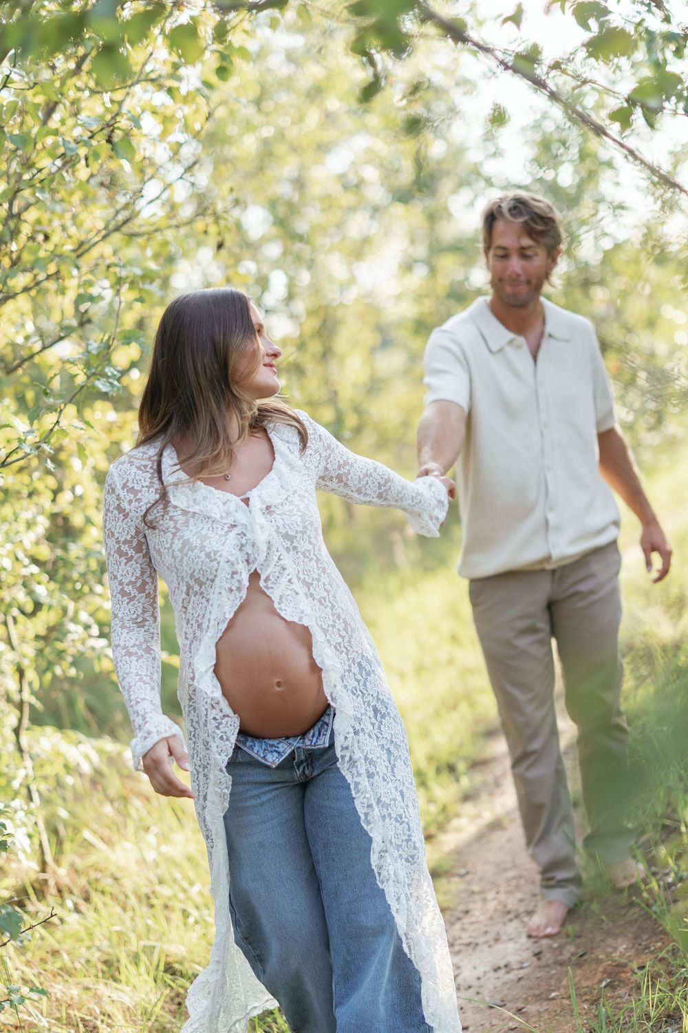 Pregnant woman in white lace cardigan and jeans walks hand-in-hand with partner on sunny forest path.