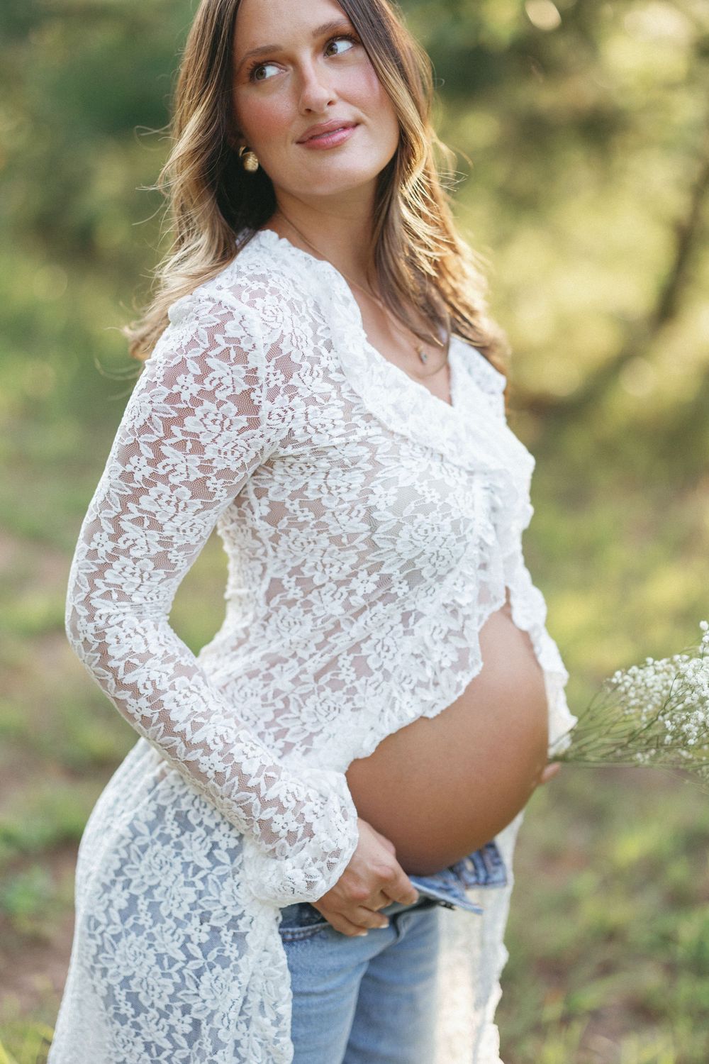 Pregnant woman in white lace cardigan and jeans cradling baby bump outdoors in sunlit green forest.