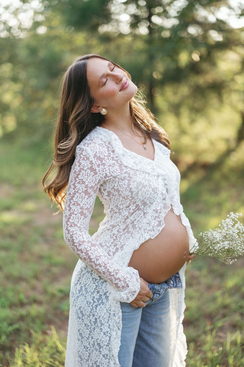 Serene pregnant woman in white lace cardigan holds wildflowers, eyes closed, glowing in golden sunlight outdoors.