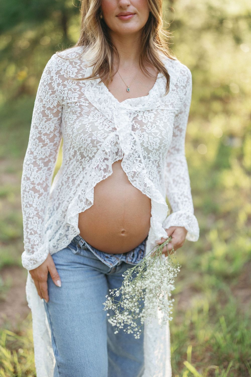 Pregnant woman in white lace top and jeans holding baby's breath flowers outdoors in natural light.