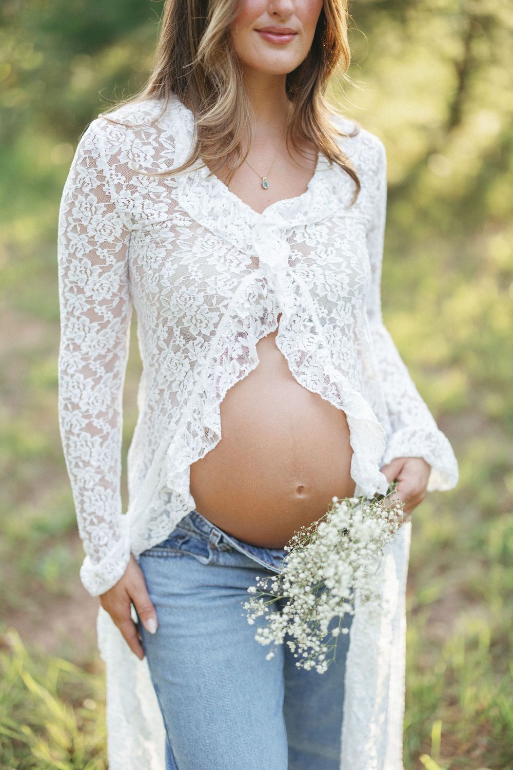 Close-up of pregnant womans bare belly holding baby's breath flowers, wearing white lace cardigan and jeans.