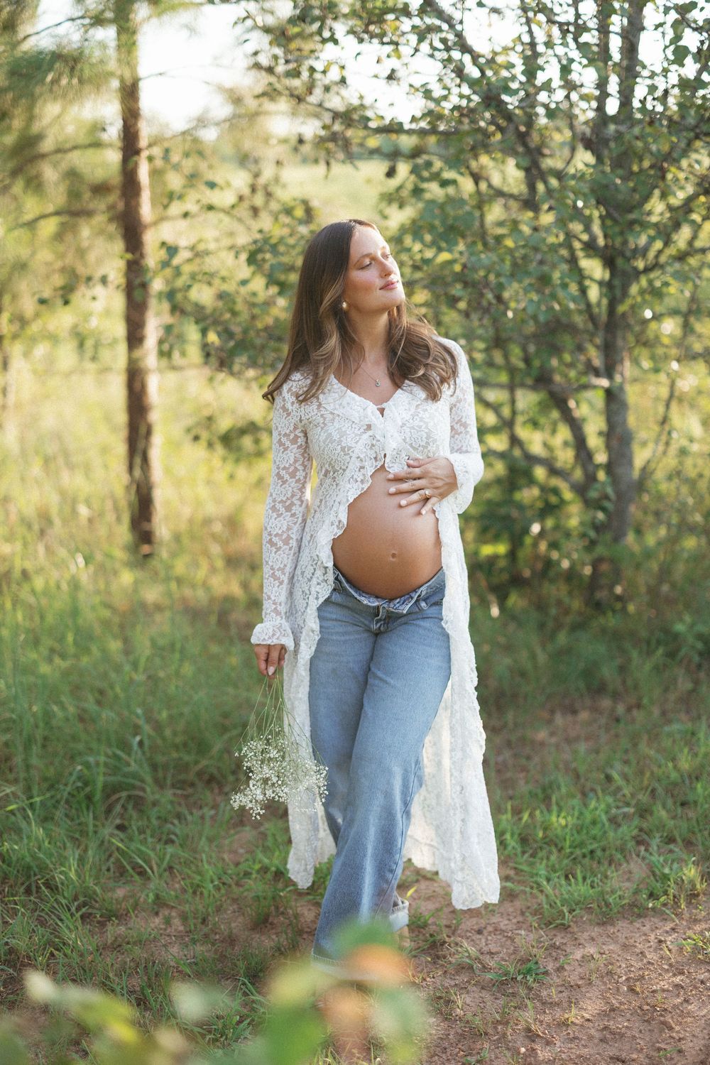 Pregnant woman in white lace cardigan and jeans holding belly outdoors in sunlit forest.