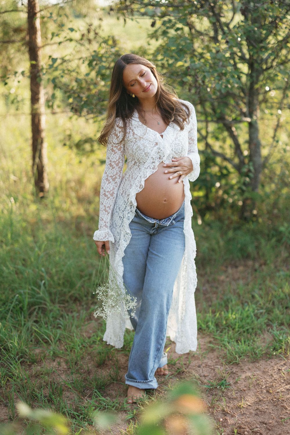 Smiling pregnant woman in lace cardigan and jeans holds belly while walking outdoors holding white wildflowers.