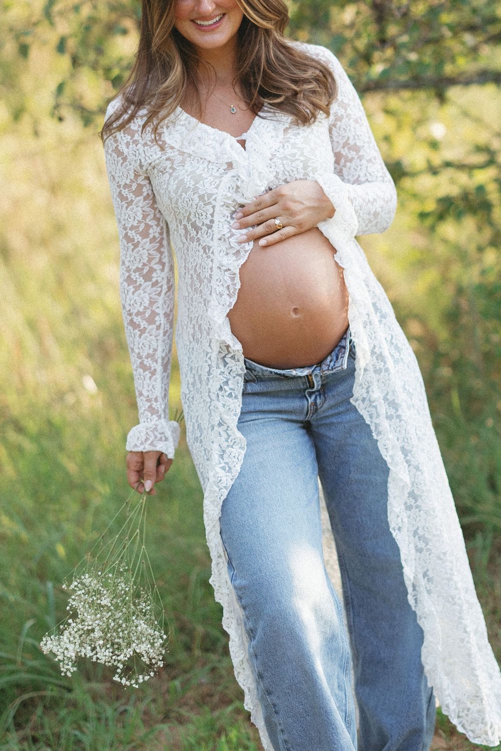 Pregnant woman in white lace cardigan and jeans holding baby's breath flowers outdoors, cradling her bare baby bump.