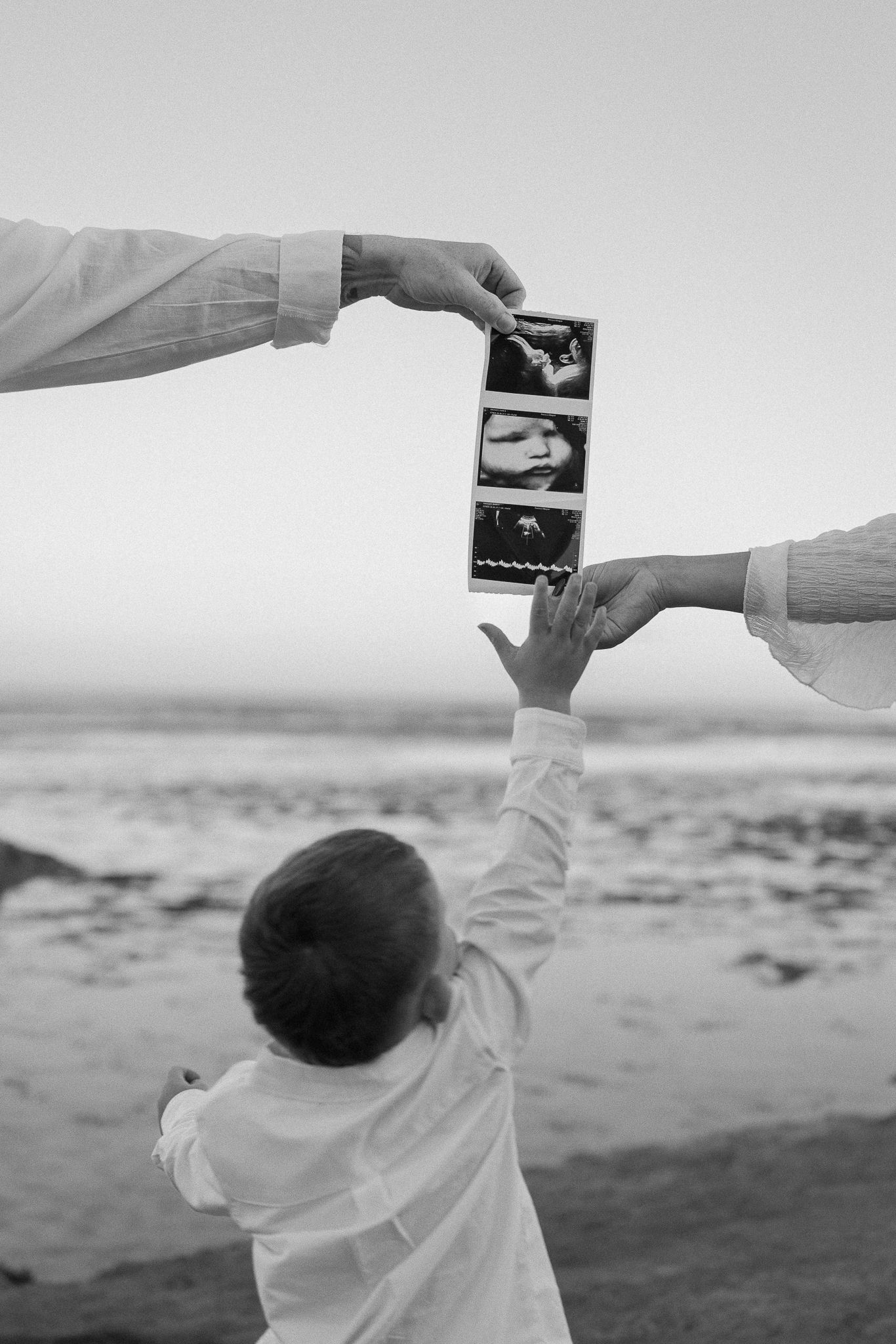 Black and white photo of child reaching for ultrasound scan held by parents on a beach.