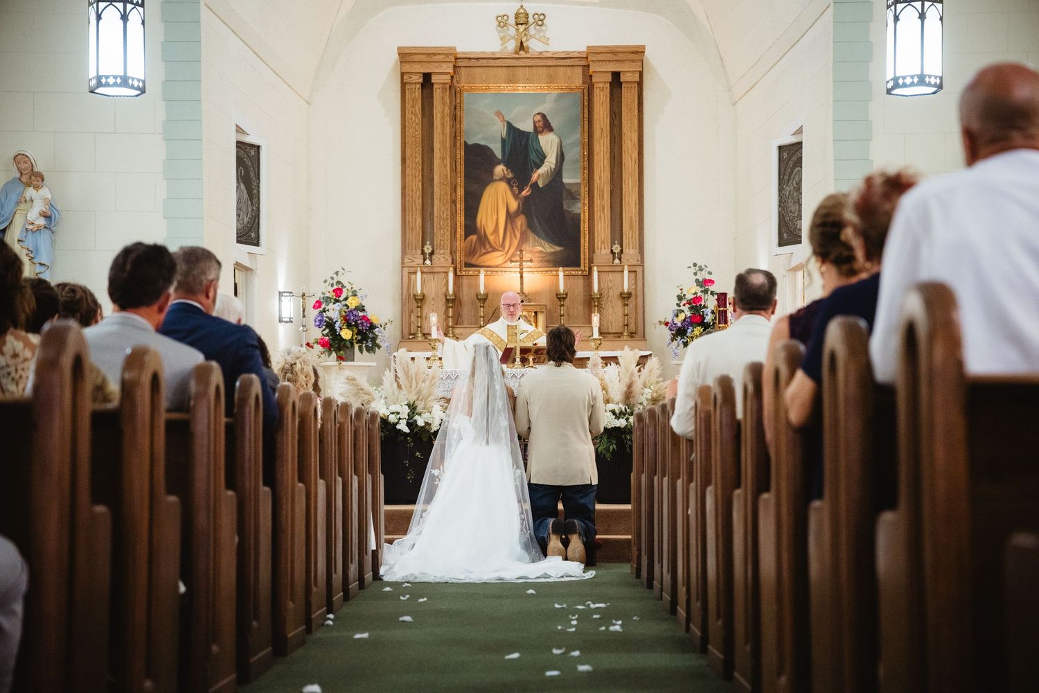 Bride and groom kneeling at church altar during Catholic wedding ceremony with flower-lined aisle and wooden pews.