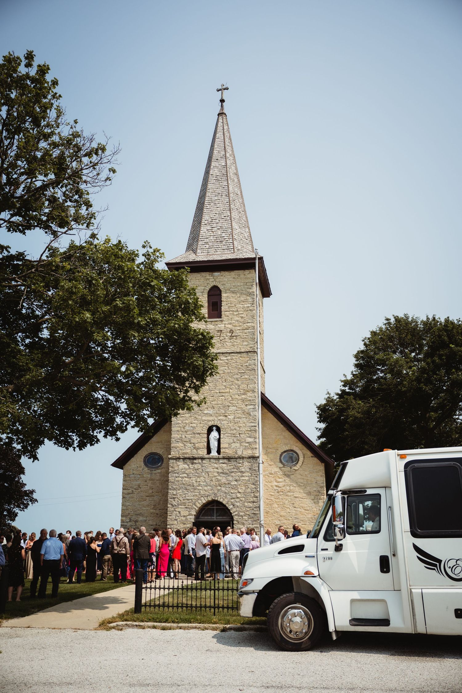 Crowd gathered outside a historic stone church with a steeple on a sunny day, with a white truck parked nearby.
