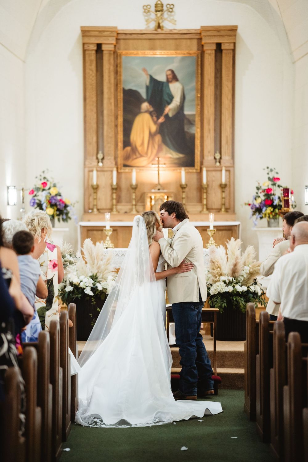 Bride and groom share first kiss at church altar during wedding ceremony, surrounded by floral arrangements and guests.