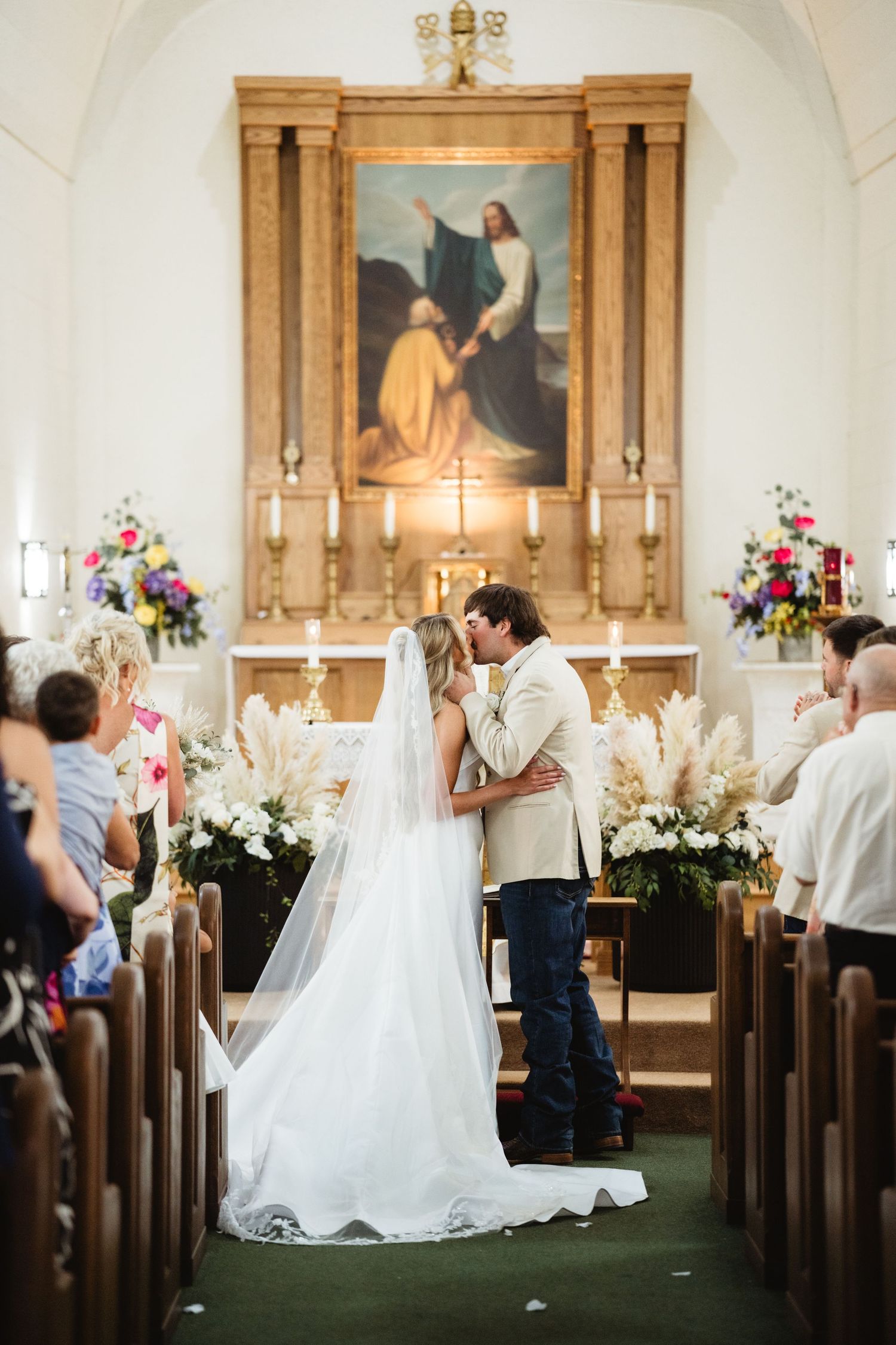 Bride and groom share first kiss at church altar during wedding ceremony, surrounded by floral arrangements and guests.