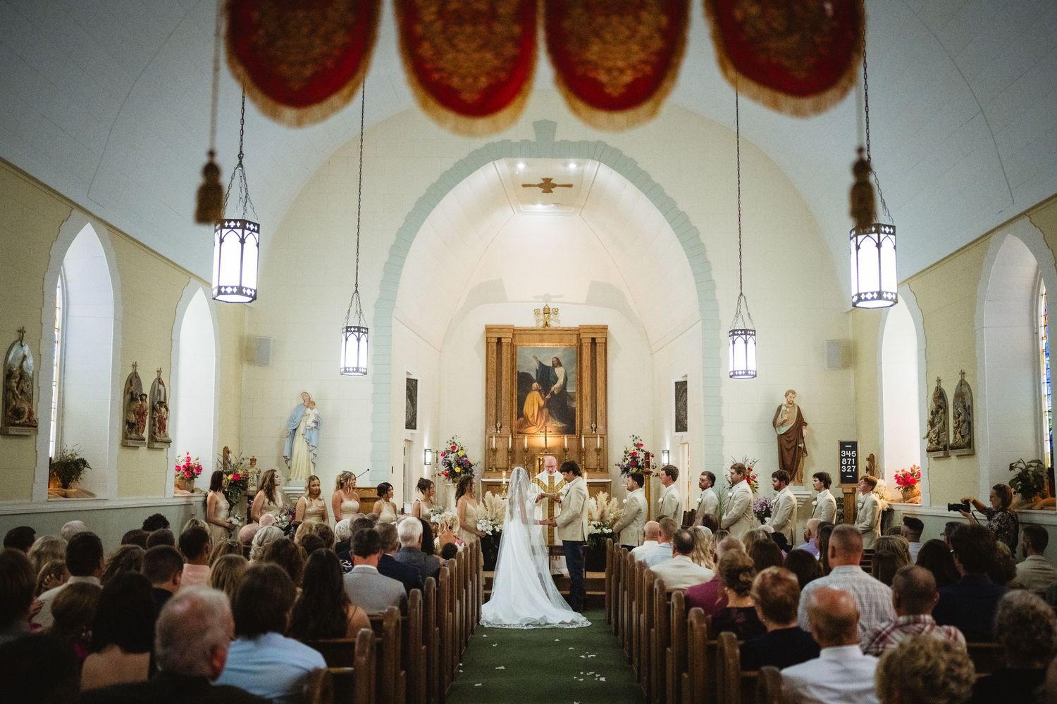 Elegant Catholic church wedding ceremony with bride and groom at altar, wooden pews filled with guests, ornate decor.