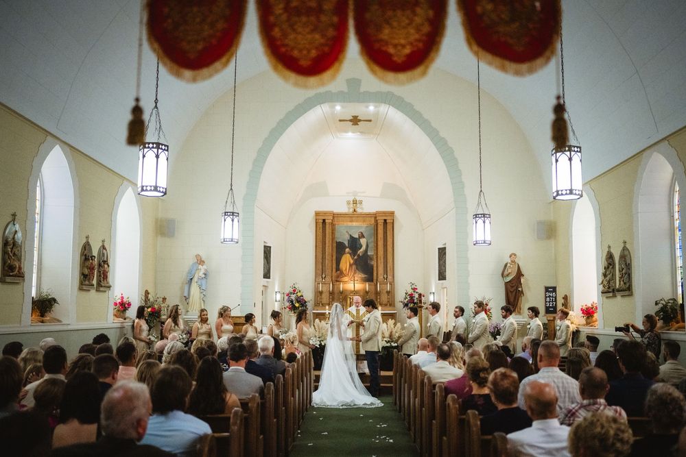 Elegant Catholic church wedding ceremony with bride and groom at altar, wooden pews filled with guests, ornate decor.