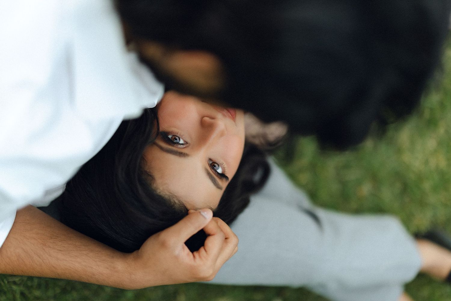 Couple lying on grass, woman looking up at man tenderly touching her face in a romantic outdoor moment.