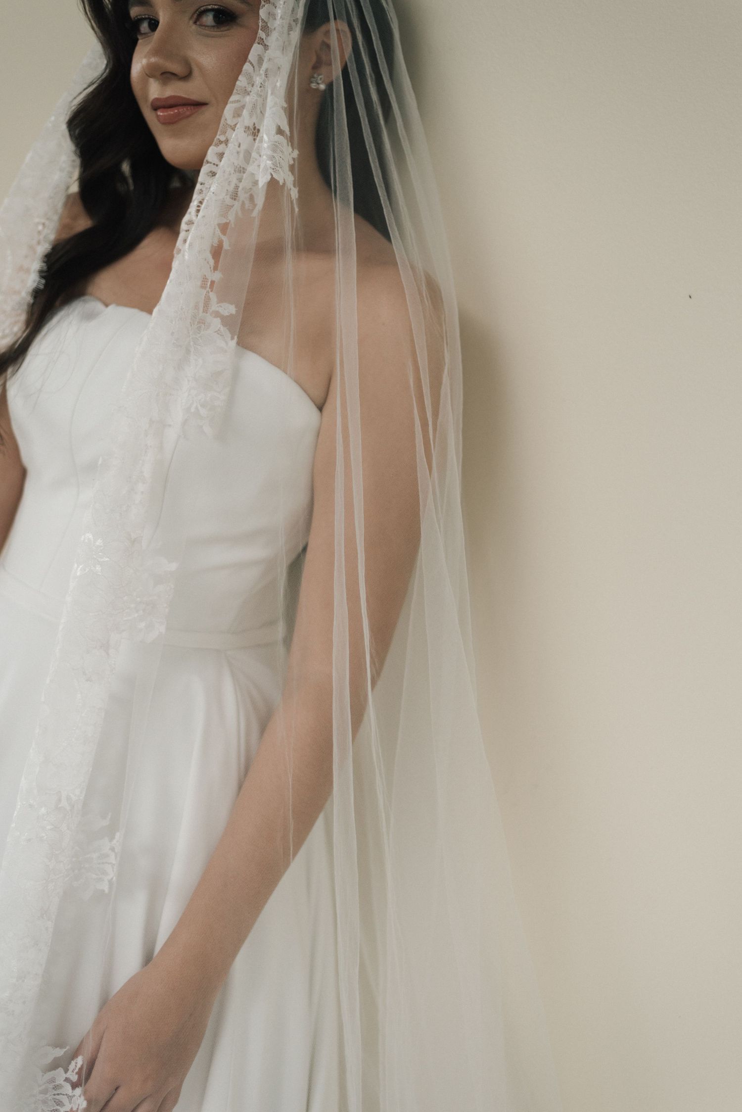 Bride in strapless white wedding gown with long lace-trimmed veil, posing elegantly against a neutral wall.