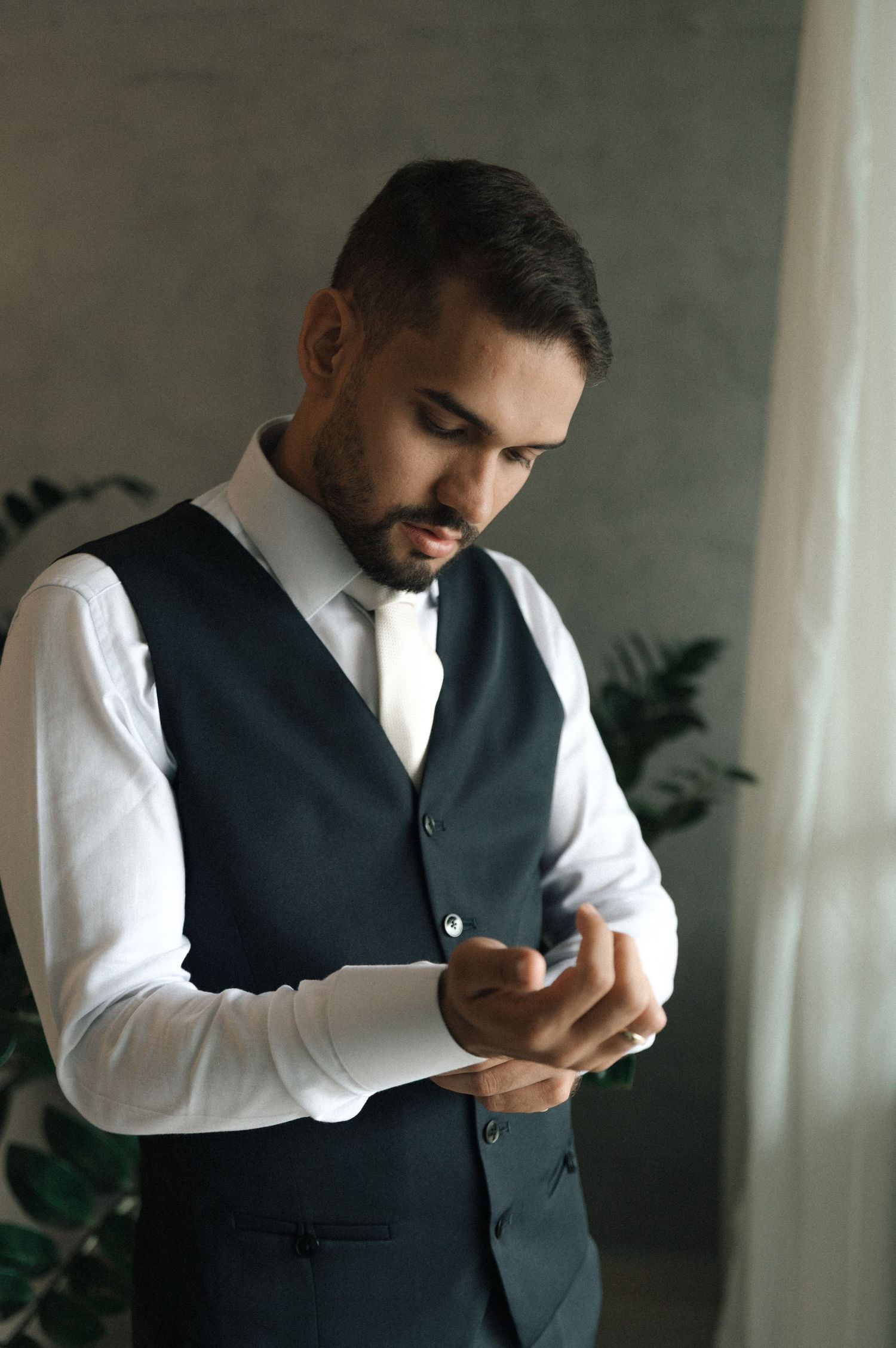 Well-dressed man in dark vest and white shirt adjusting cufflinks indoors.