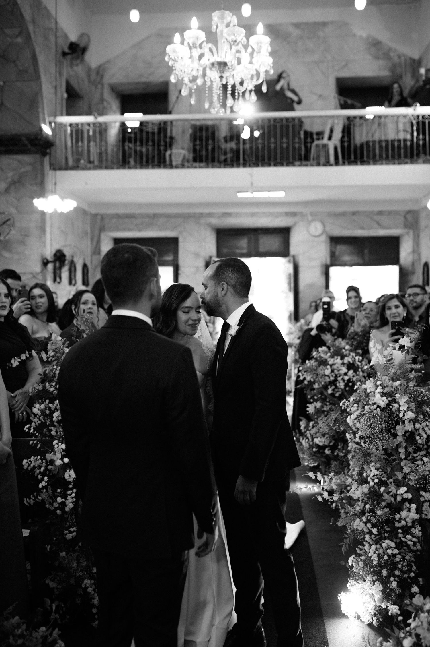 Black and white wedding ceremony photo of couple sharing a kiss at the altar, surrounded by floral arrangements and guests.