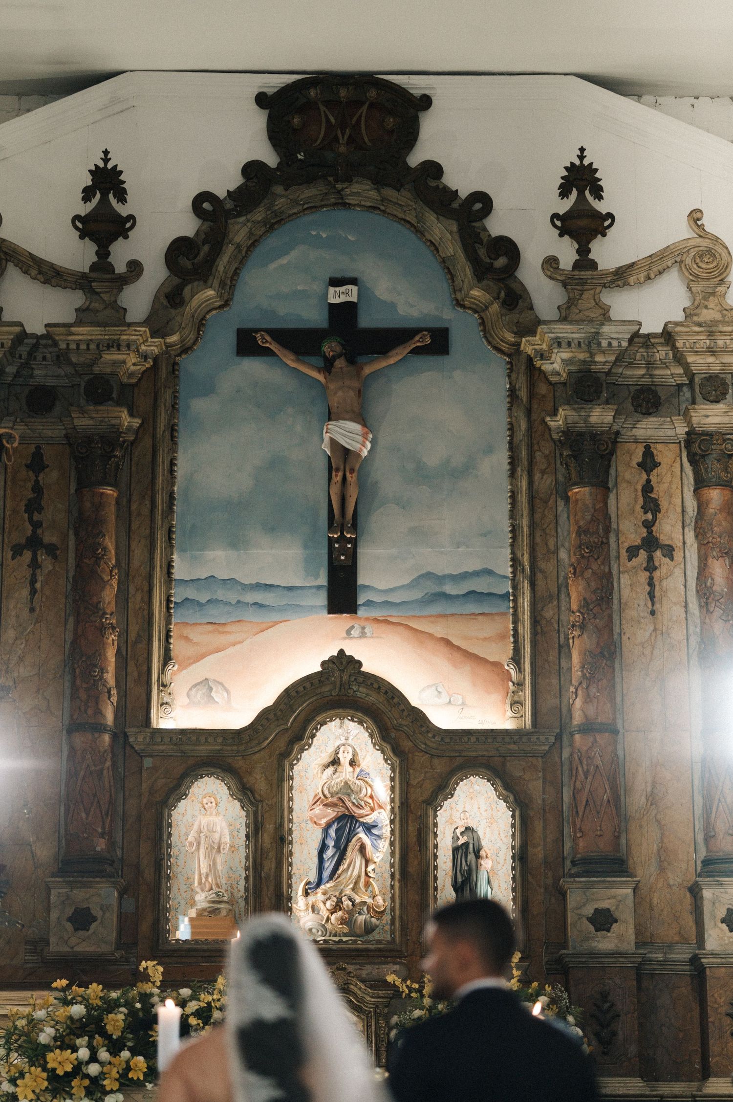 Couple stands before ornate church altar with crucifix painting during a wedding ceremony.