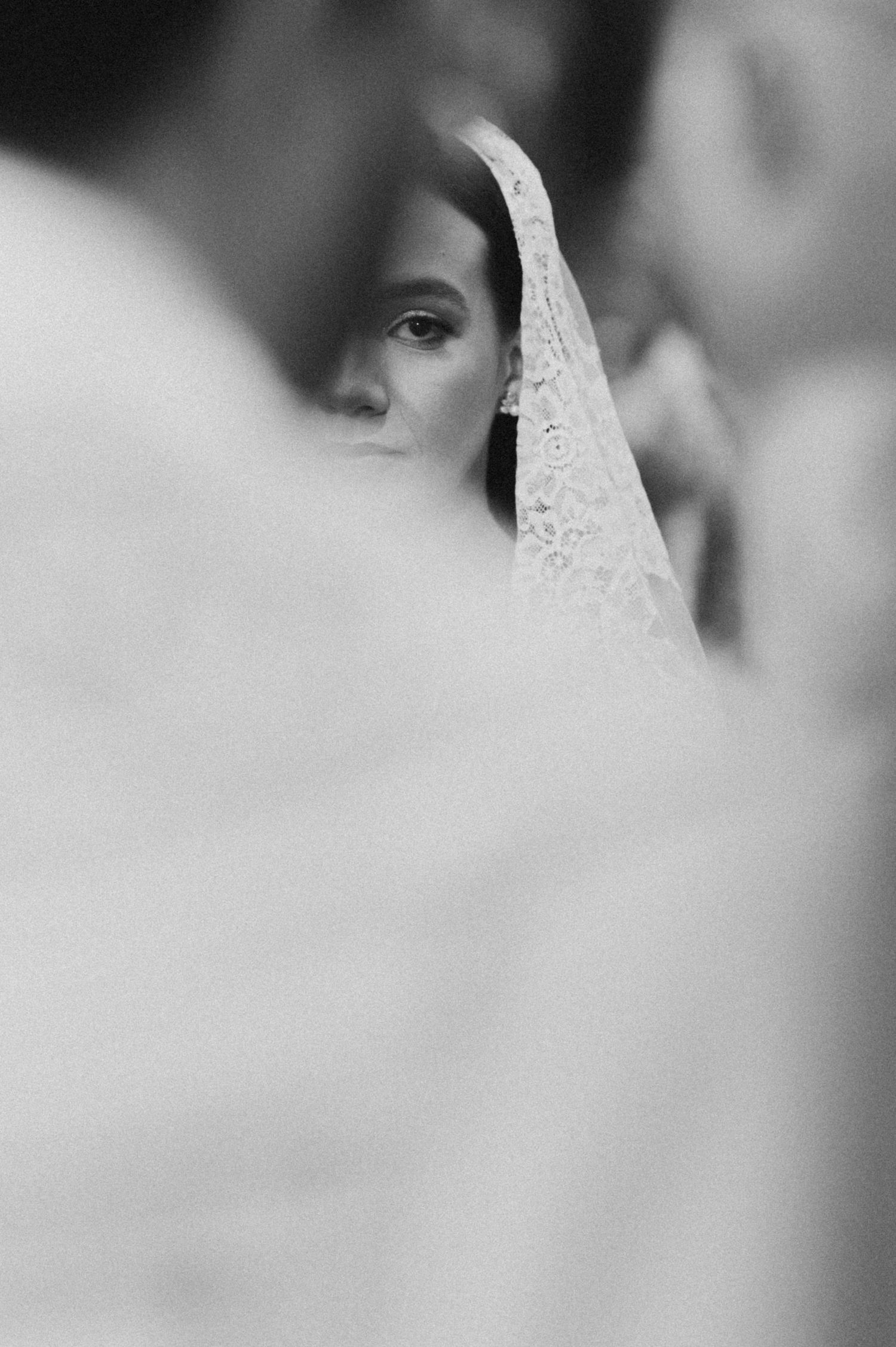 Black and white wedding photo of a bride in lace veil, partially framed by blurred figures in the foreground.