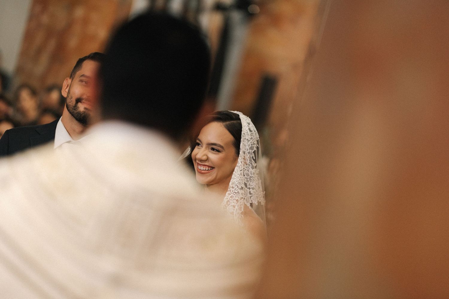 Smiling bride in lace veil during wedding ceremony, groom beside her, officiant in foreground.