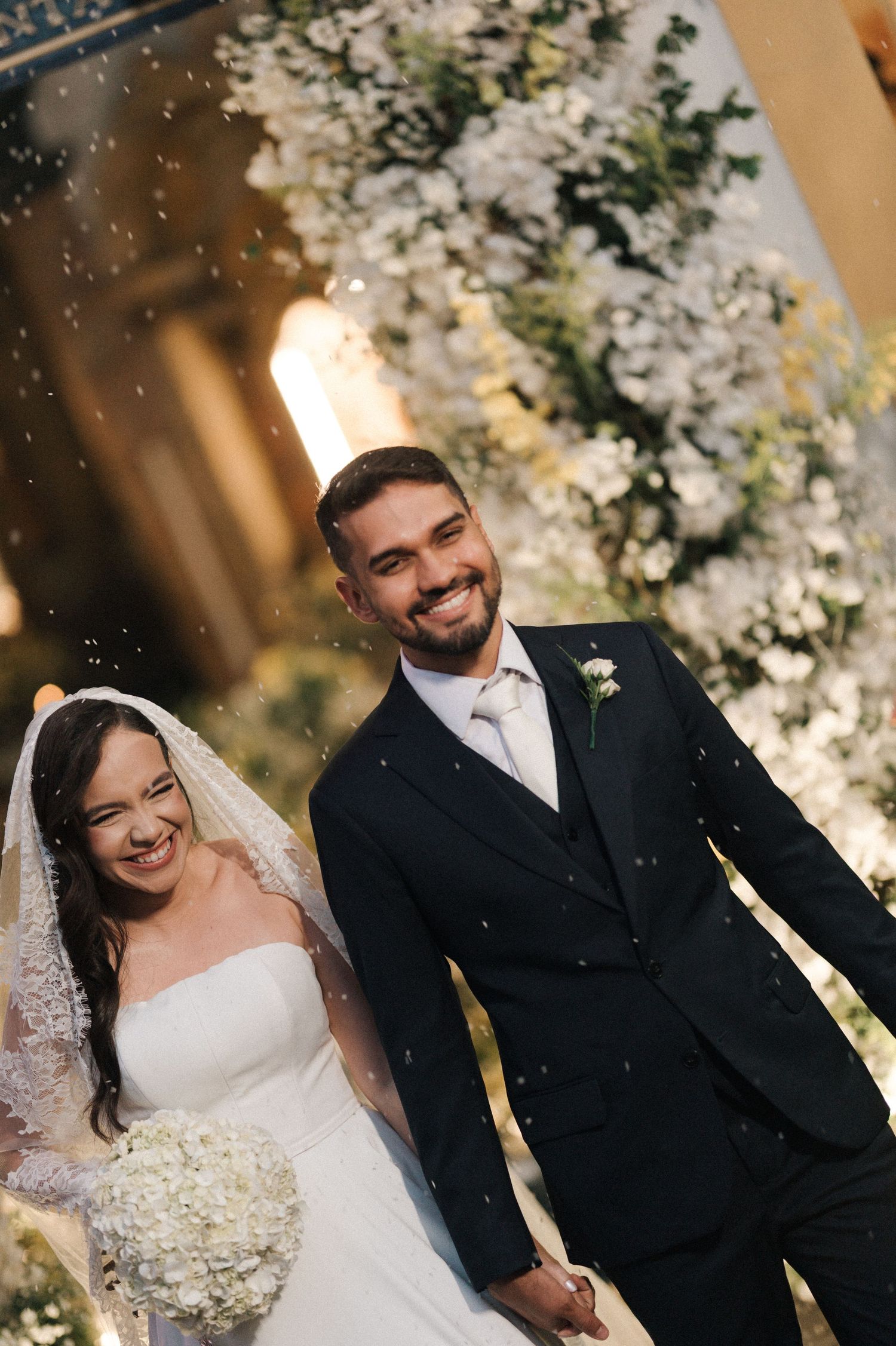 Smiling bride and groom walking together after wedding ceremony, bride holding white bouquet, groom in black suit.