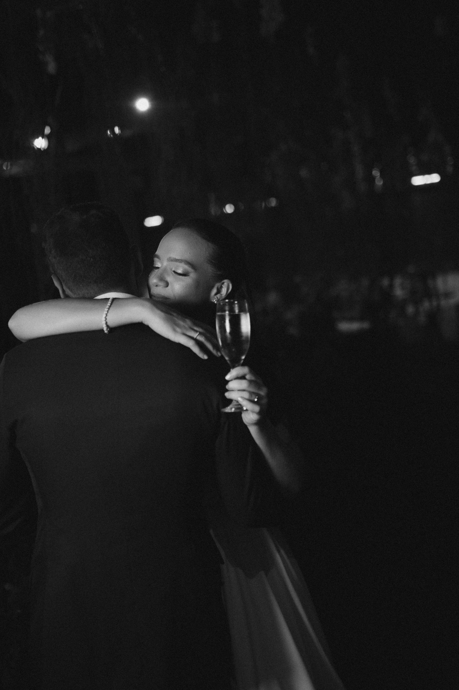 Black and white photo of a bride hugging her groom while holding a champagne glass at their wedding reception.