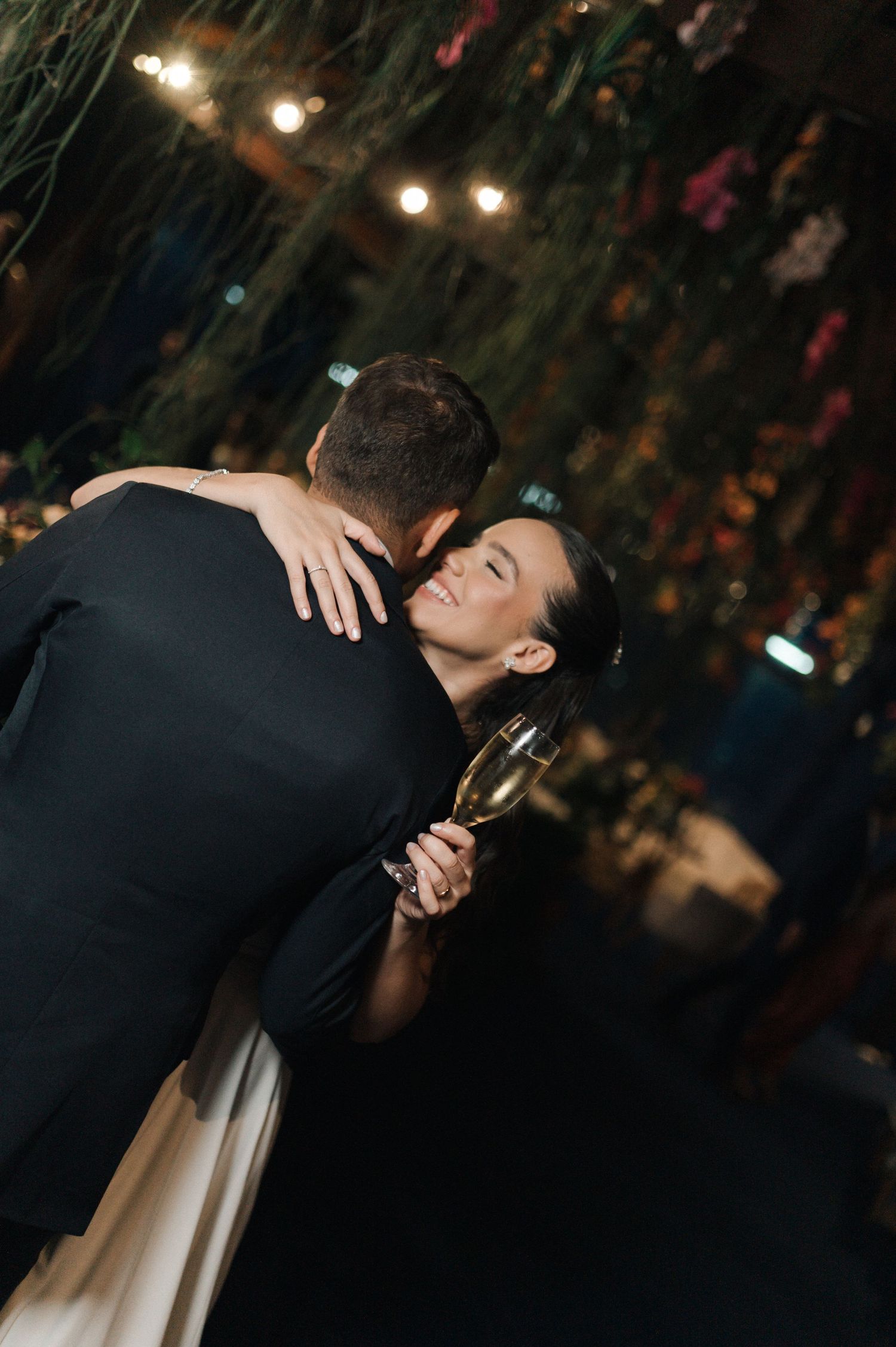Couple sharing a romantic embrace at a wedding reception, woman holding champagne glass under warm string lights.