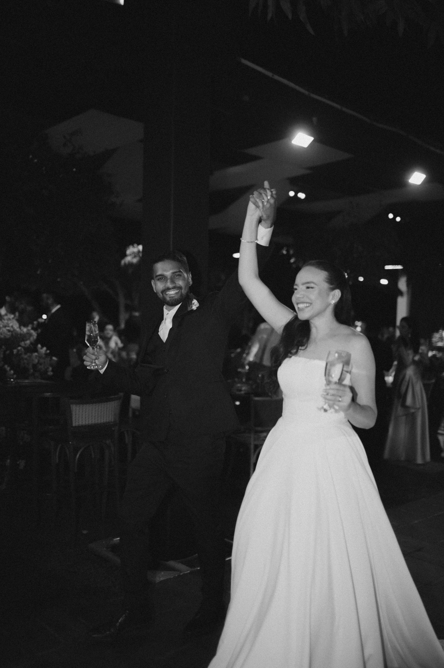 Black and white photo of a joyful bride and groom raising glasses at their wedding reception at night.