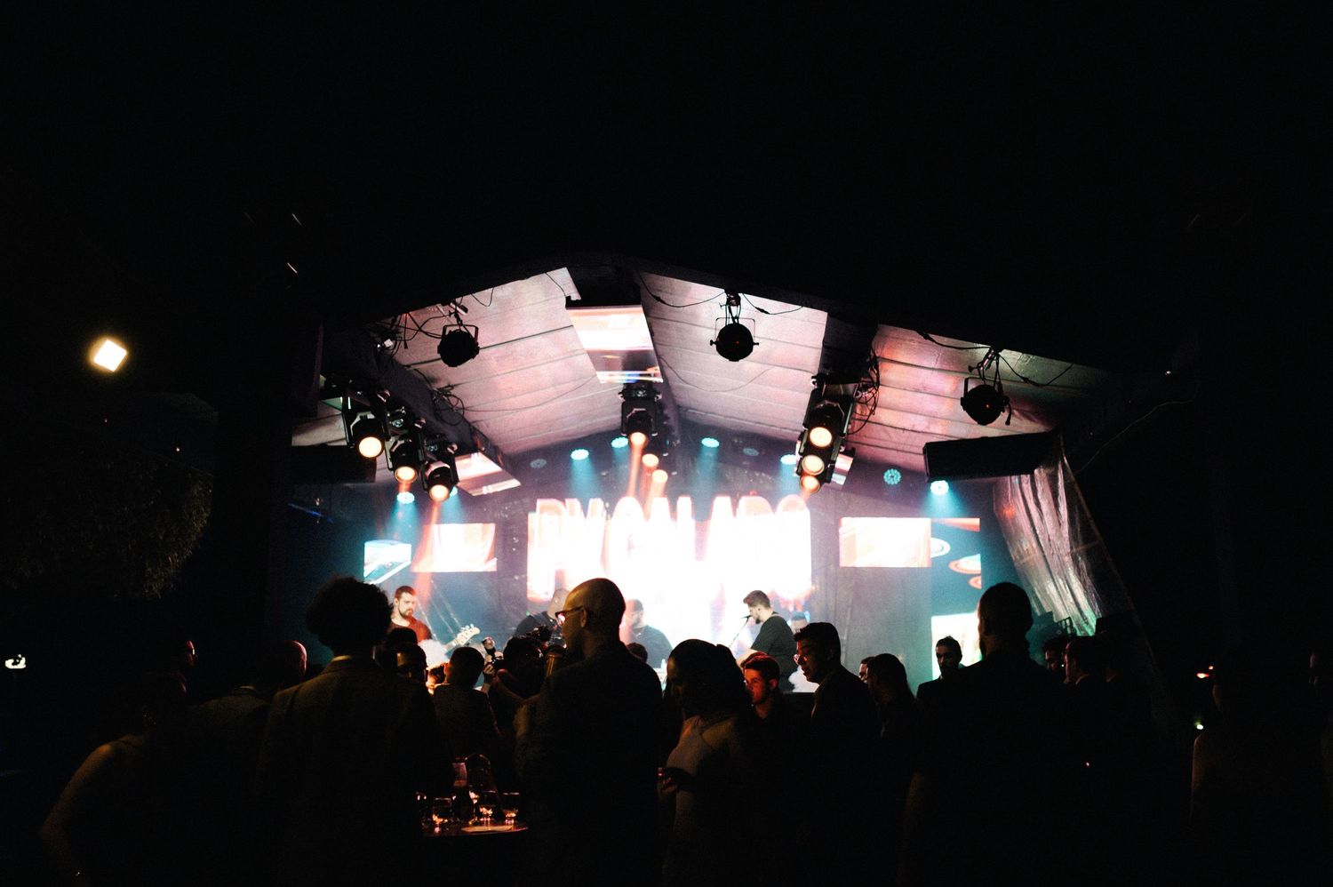 Crowd watching a live band perform on a brightly lit stage at an indoor concert venue at night.