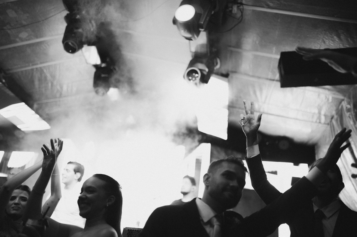 Black and white photo of crowd at a party with smoke and raised hands under bright stage lights.
