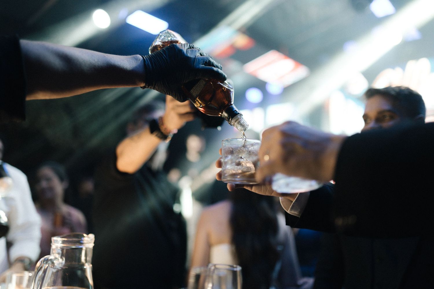 Bartender pouring whiskey into shot glasses at a lively nightclub bar event with guests in background.