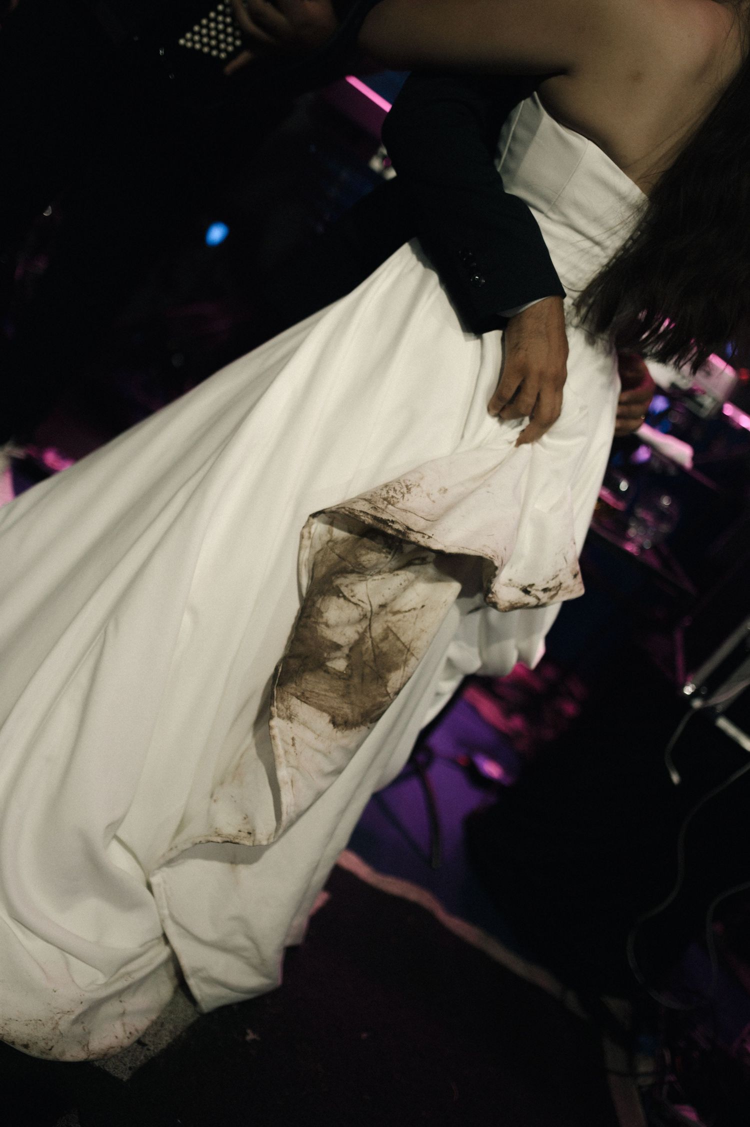 Person wearing a dirty white wedding gown with mud stains on stage under colorful lights.