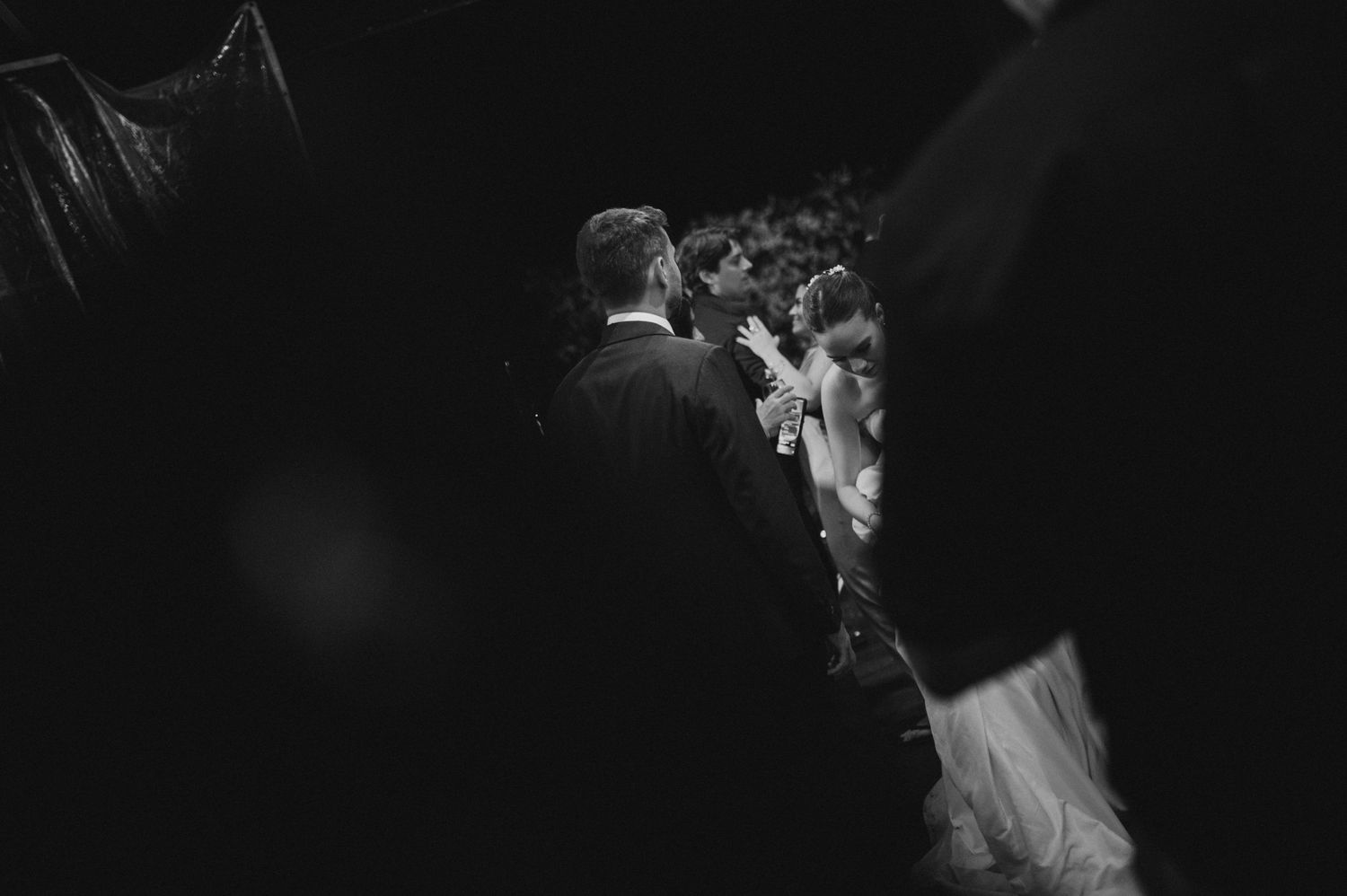 Black and white wedding photo of bride and groom sharing a moment surrounded by guests in elegant attire.
