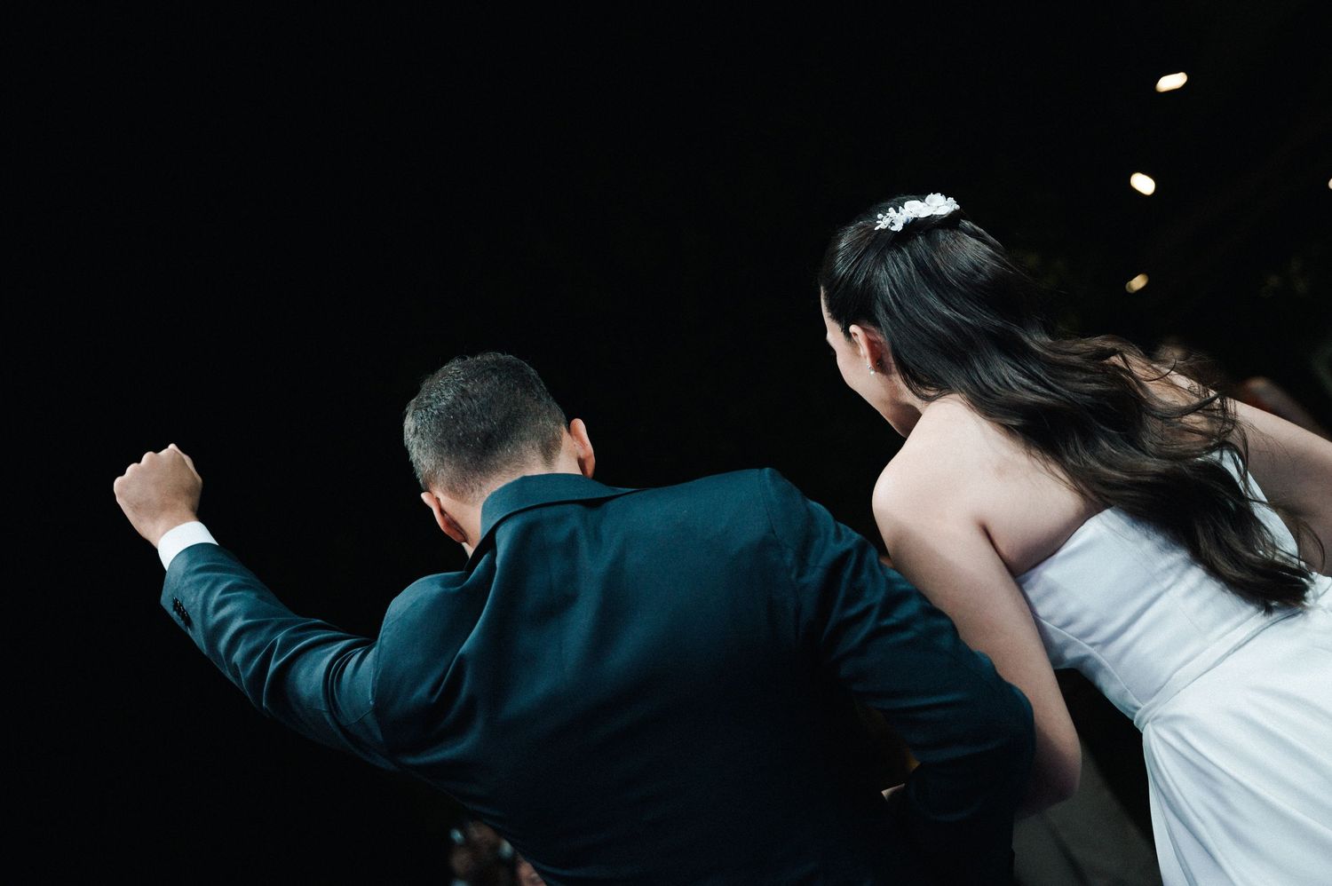 Bride and groom walking away, man in teal suit raising fist, woman in white wedding dress with floral hair accessory.