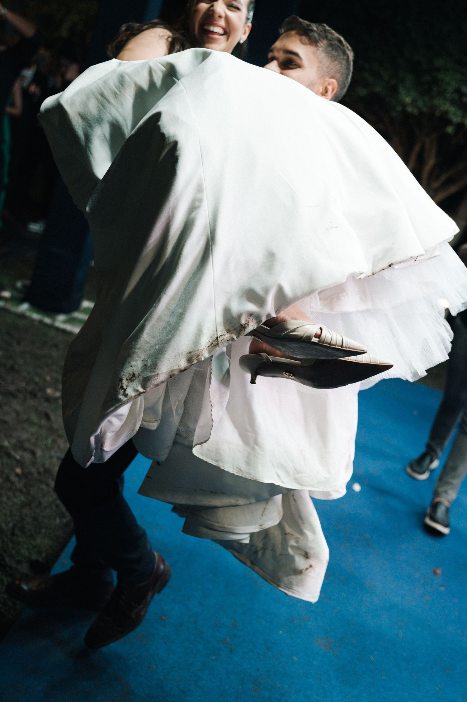 Groom carrying bride in white wedding dress on blue carpet at outdoor evening celebration.