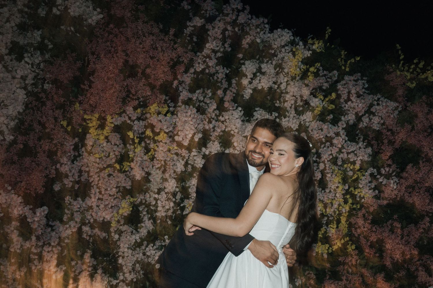 Bride and groom embracing romantically in front of blooming pink floral backdrop at night wedding celebration.
