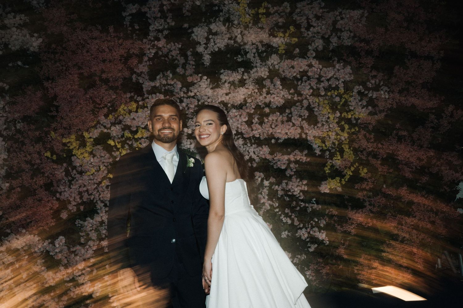 Bride and groom smiling together in front of a floral wall backdrop at their wedding reception.