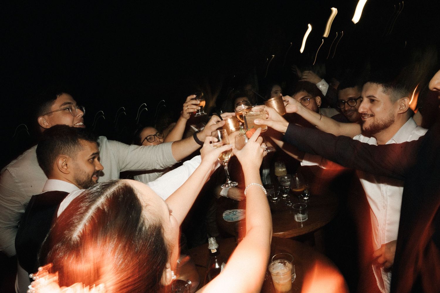 Group of friends raising glasses in a celebratory toast at a dark, lively party event.