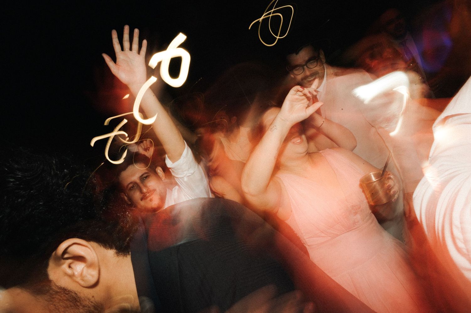 Blurred motion photo of people dancing at a party with light trails and bokeh effects in dark setting.