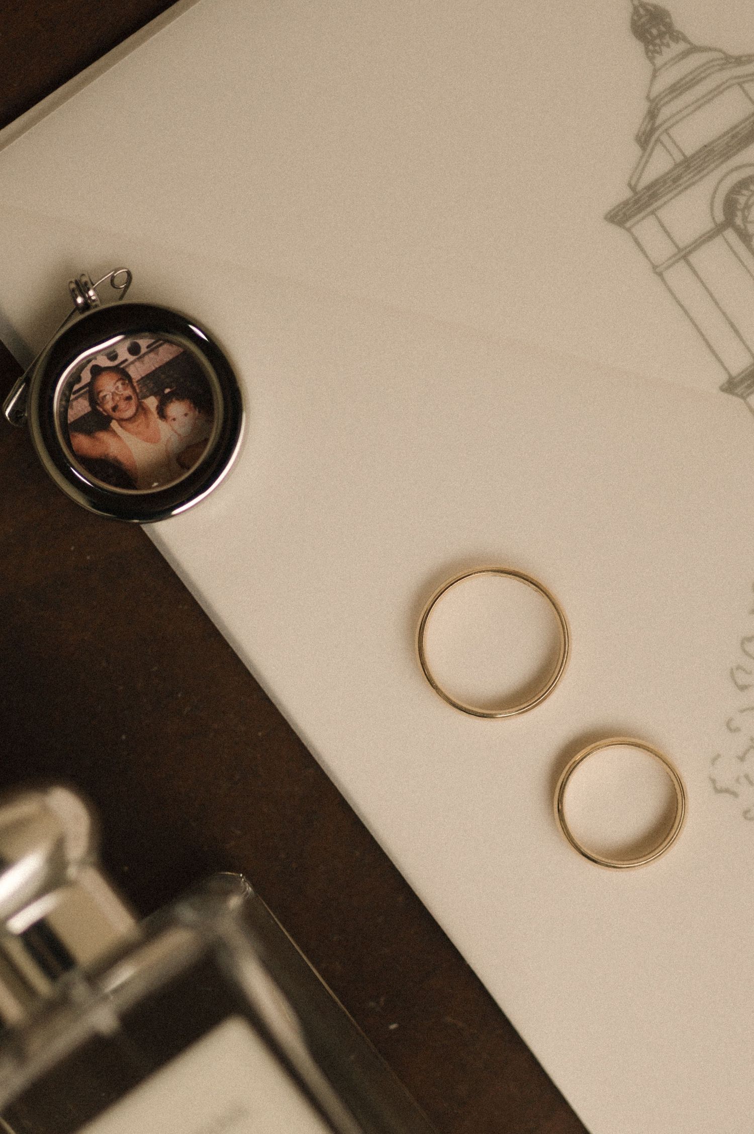 Two gold wedding bands and a locket with a couple's photo rest on a white wedding invitation card.