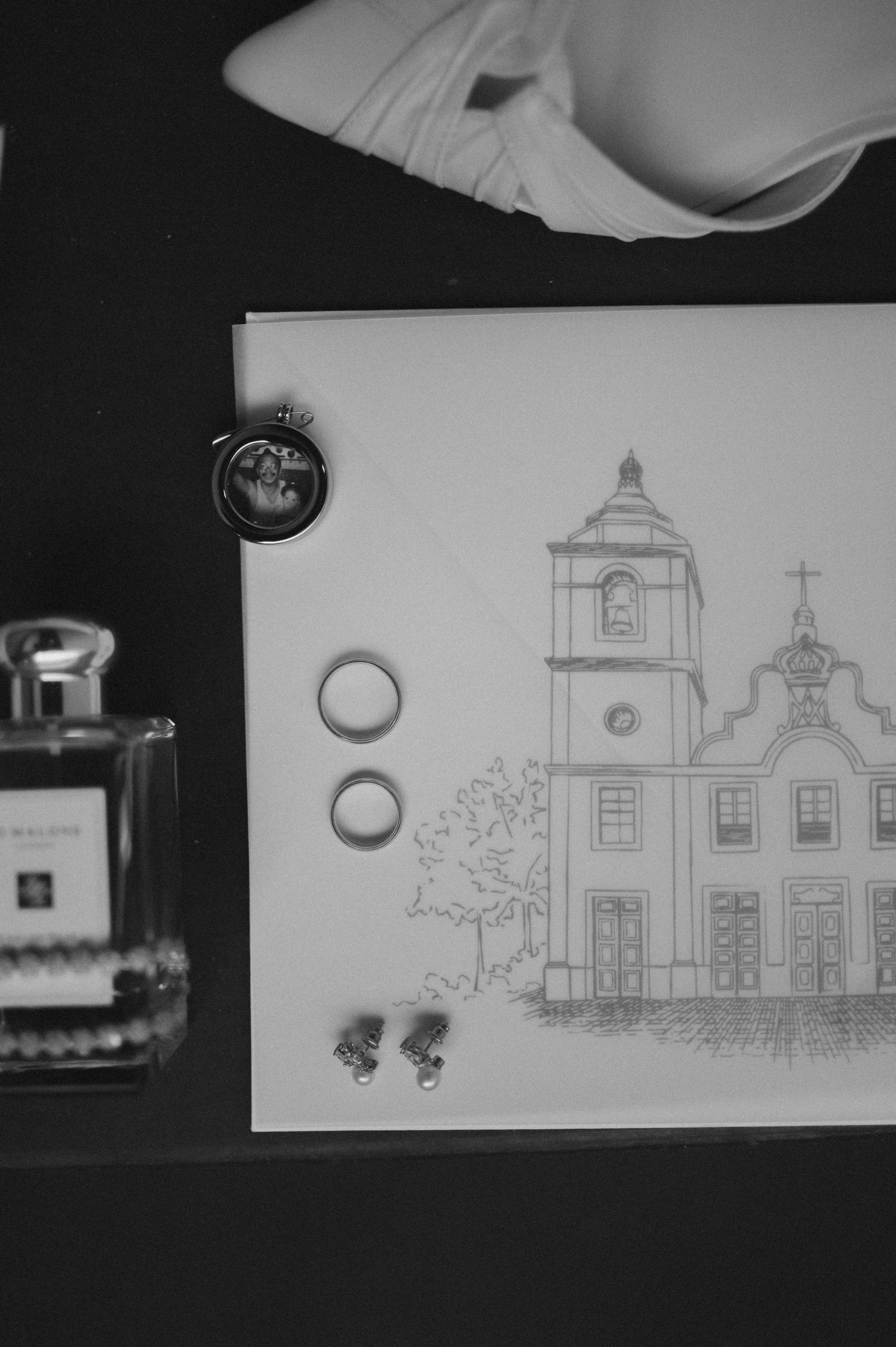Black and white wedding flat lay with two gold rings, earrings, perfume bottle, and a sketched church illustration on white card.