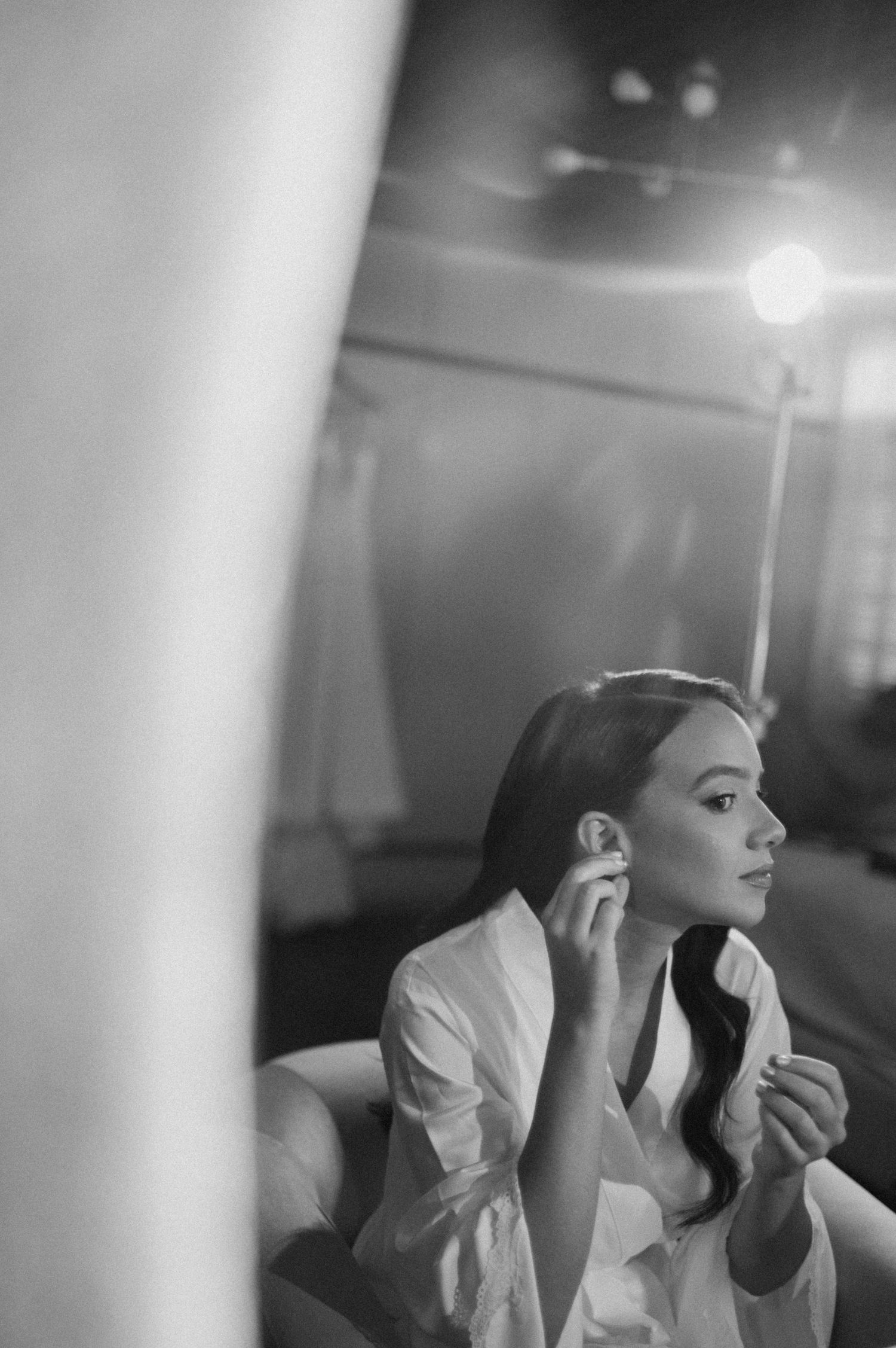 Black and white photo of a woman in a white shirt putting on an earring while seated in a softly lit room.