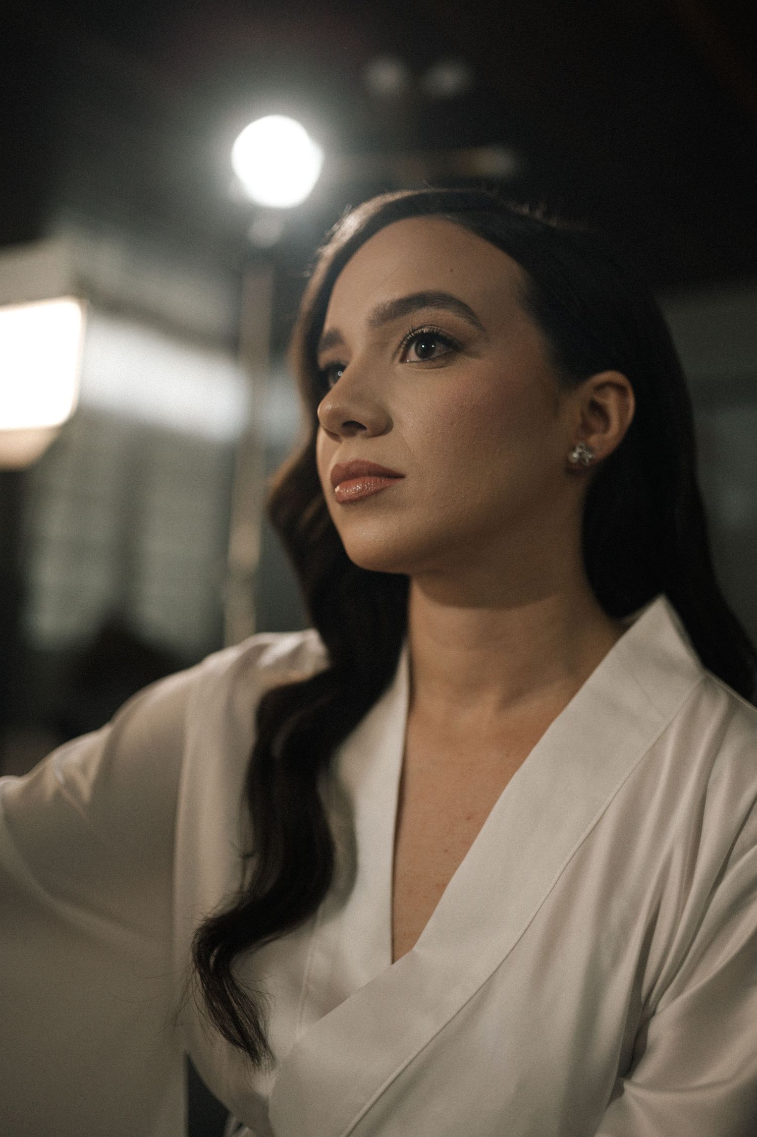 Young woman with long dark hair in white wrap top gazes upward under studio lighting.